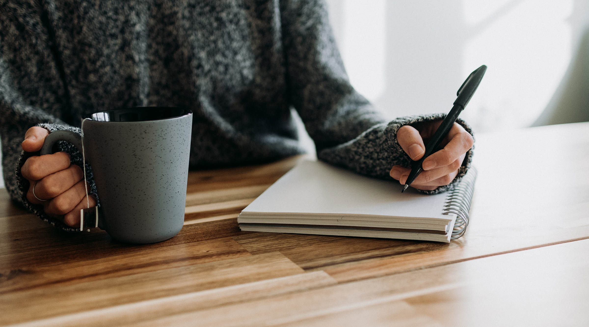 A person holding a pen, about to write in a notebook and sitting at a table. In their other hand is a cup of tea. Photo by Kelly Sikkema, via Unsplash