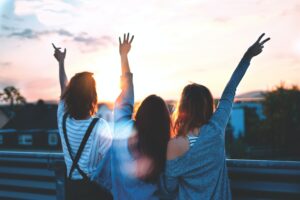 Three young people standing together facing sunset with arms raised in celebration