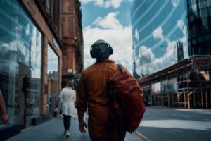 Student wearing over-ear headhpones with rucksack on right shoulder walking down London street looking at blue sky with clouds