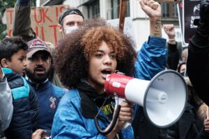 Woman holding megaphone and raising arm in protest, flanked by demonstrators with cardboard signs