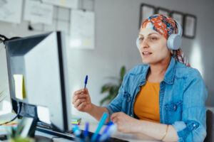 Woman with cancer in headscarf working at desktop computer with headphones