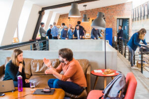 Colleagues in open plan office space having informal discussion on sofa while others gather and share breakfast