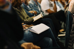 A row of people sitting at a conference. In the foreground, a person sits with a notebook, pen and smartphone on their lap.