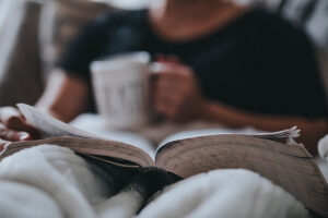 A person sitting under a blanket, holding a coffee mug and reading a book