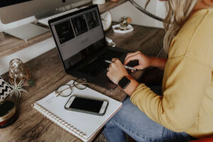Person sitting at a desk, using a laptop, with a notebook and phone next to them - photo by Corinne Kutz, via Unsplash