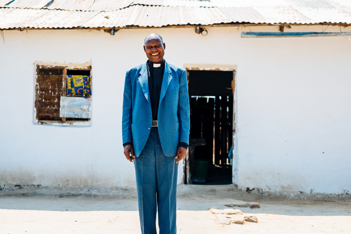 A man, Canon Atanasi, dressed in pastor's clothes, standing outside a building in Tanzania.