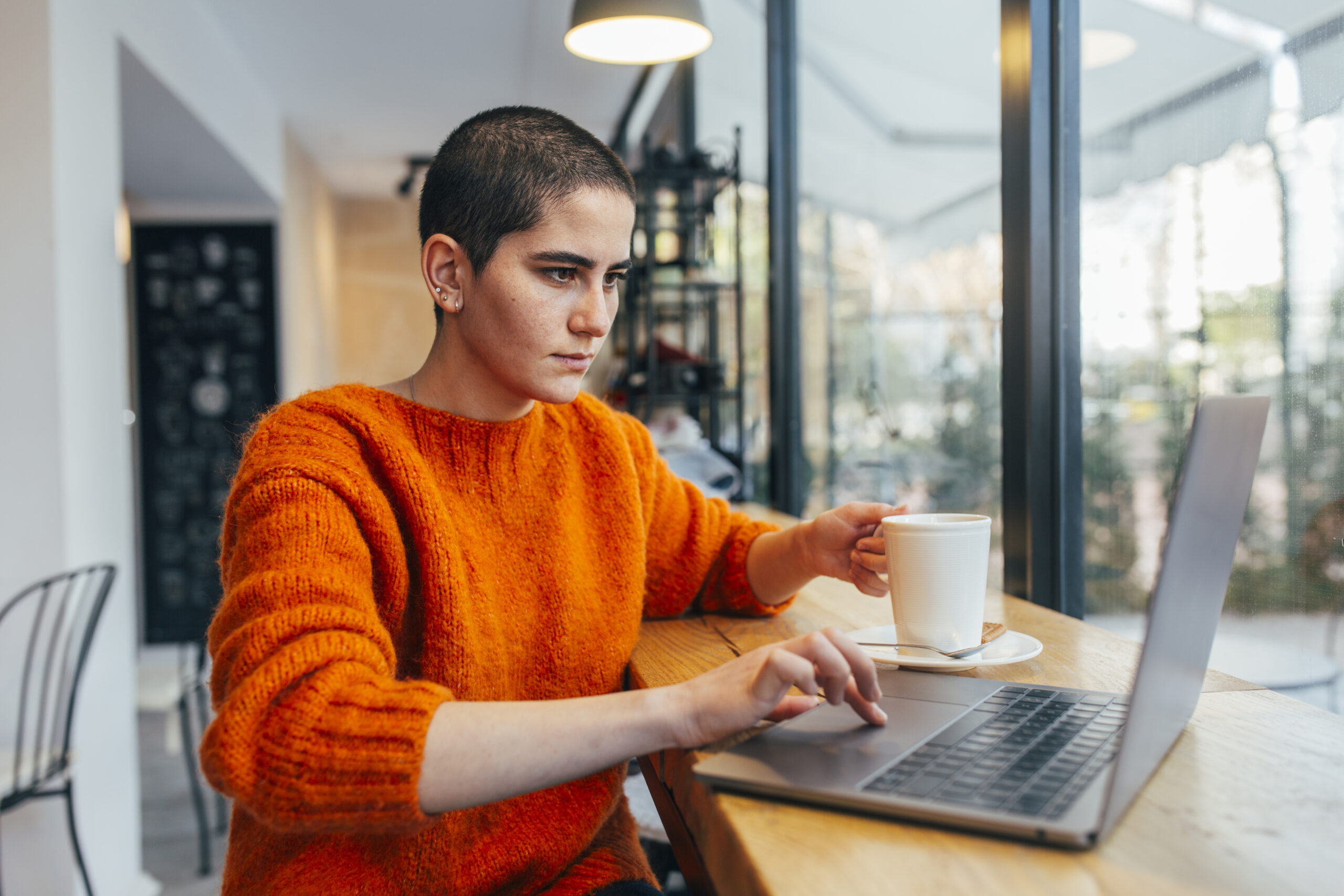 Young bald woman sitting at desk working on laptop with a coffee in cafe.