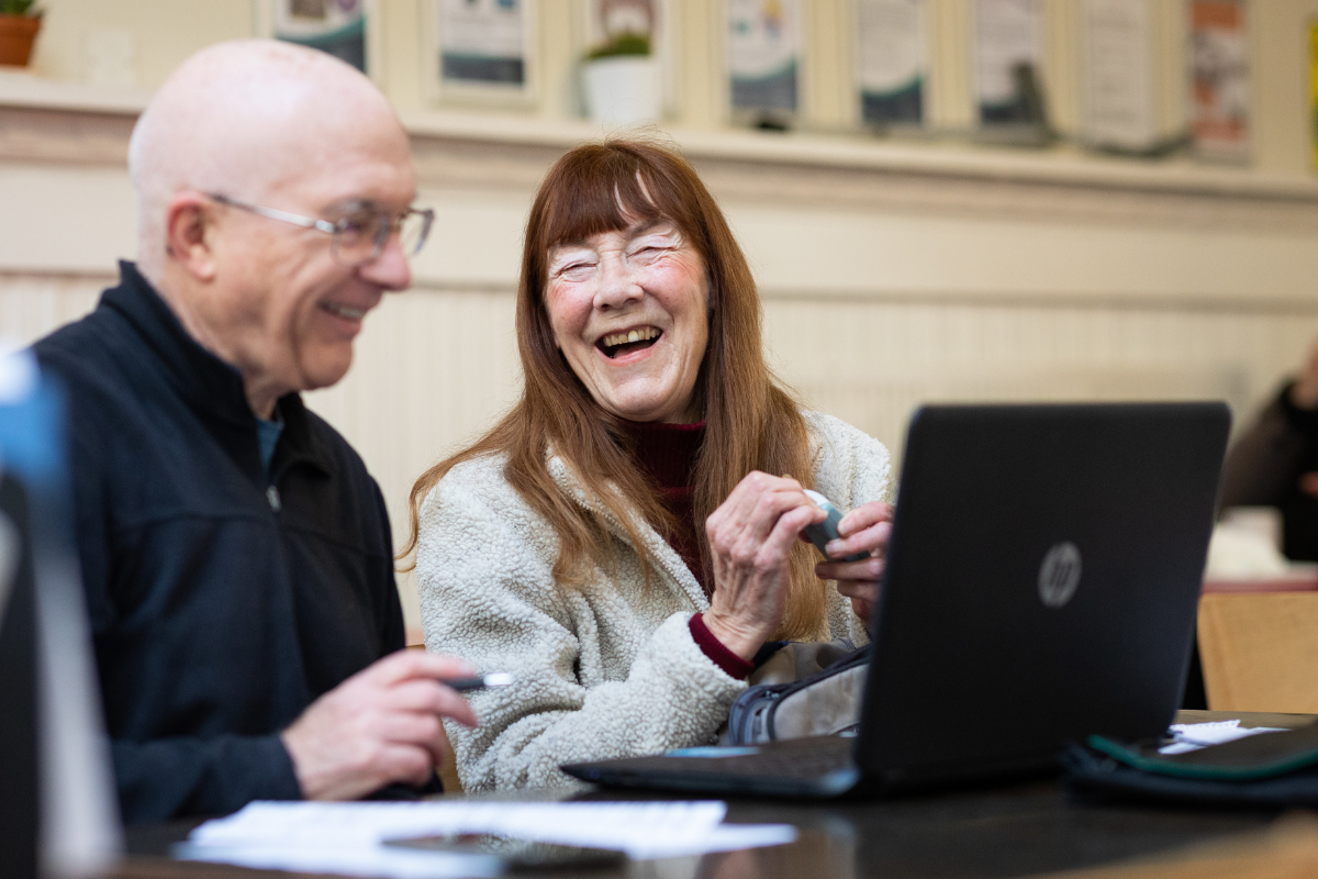 A photo of two people, an older man and woman, sat at a computer - both are looking at the screen and laughing.