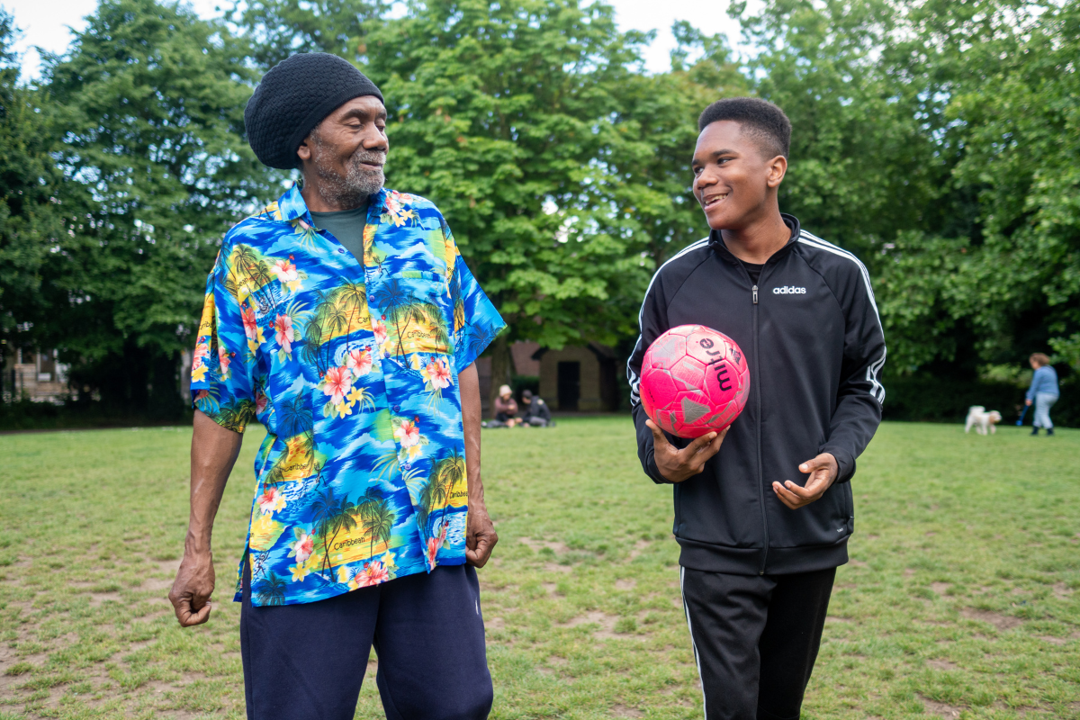 An older man and a younger man or child outside in a green area. The younger person is carrying a colourful ball.